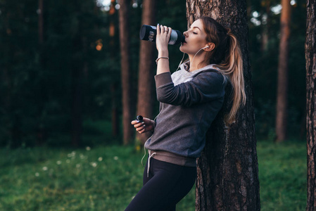 女性慢跑的恢复在密集的锻炼之后站立在树附近饮用水, 佩带耳机在公园
