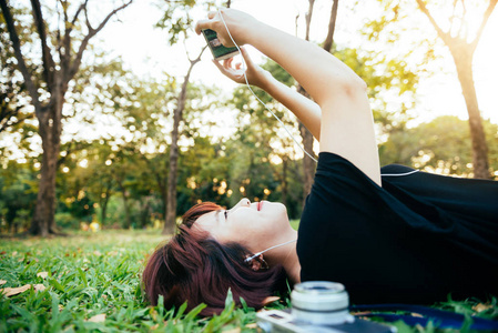 年轻的亚裔女子躺在草地上, 在公园里听音乐, 心情很冷。在草地上休息的年轻女子, 她的相机旁边。户外活动在公园概念