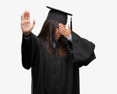 年轻的西班牙裔妇女穿着毕业帽和制服覆盖眼睛用手和做停止手势与悲伤和恐惧的表达。尴尬和消极概念