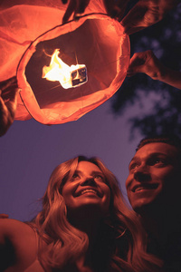 低角度观看微笑的年轻夫妇晚上在河边海滩上发射天花灯