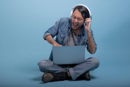 年轻的大胡子男子坐在地板上使用计算机在蓝色背景。非常兴奋的亚洲青年时髦使用笔记本电脑听音乐, 全身射击。年轻一代时髦的工作理念
