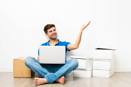穿着蓝色衬衣的年轻人坐在他 cardbox 旁边的地板上, 在网上看公寓, mooving 去新房。在白色背景上