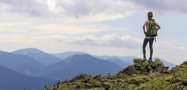 年轻苗条的女孩与背包站立在岩石山顶对明亮的蓝色清晨天空享受雾山脉全景的背面视图。旅游旅行和健康的生活方式概念