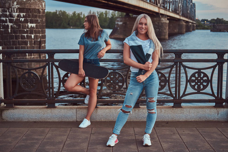 两个美丽的时髦女孩站在与滑板的桥梁