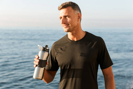英俊强壮成熟运动员的照片站在沙滩上喝瓶装水