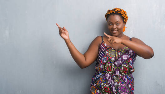 年轻的非洲裔美国妇女在灰色的垃圾墙上穿着橙色辫子微笑着, 看着相机指着两只手和手指的侧面