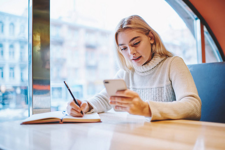 十几岁的女孩阅读智能手机上的新信息, 而在咖啡馆内部学习准备作业任务, 年轻女性时髦的检查通知蜂窝状创建文章为博客出版物