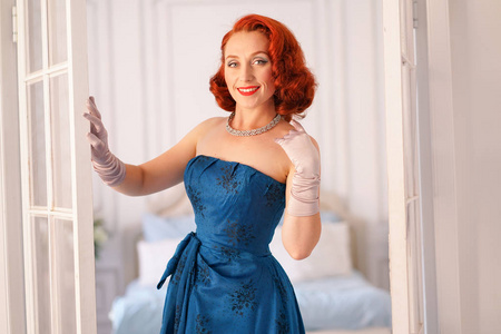 一个豪华的别针女士穿着蓝色的复古礼服站在她的卧室门口, 并邀请你进去