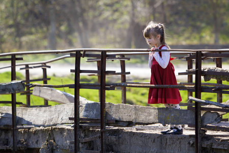 漂亮的小金发女孩穿着漂亮的红色连衣裙独自站在旧的水泥桥上, 倚着木栏杆, 目不转睛地看着温暖的阳光明媚的一天。儿童好奇心和幻想概
