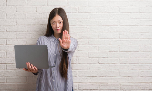 年轻的中国妇女在砖墙使用手提电脑与张开手做停止标志以严肃和自信的表达, 防御手势