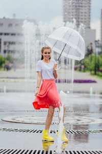 年轻漂亮的女孩与两个辫子在黄色靴子和透明的雨伞站在喷泉附近。城市的雨天