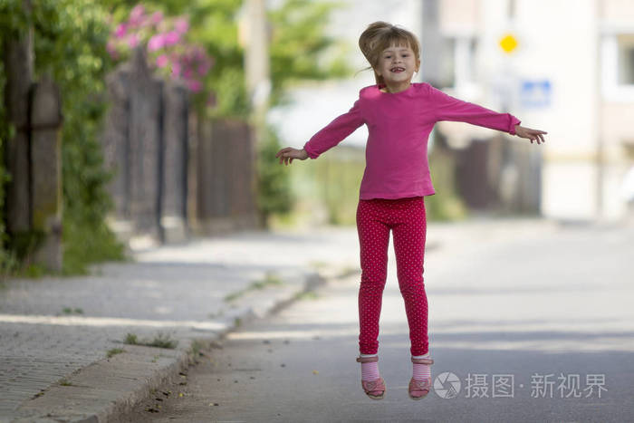 可爱的小滑稽微笑无牙的女孩在粉红色休闲服装与长的金发小马尾巴跳跃和在模糊明亮的阳光明媚的街道户外背景的乐趣。快乐的粗心童年概念