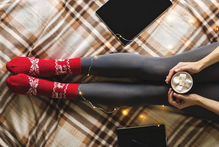 妇女的手和脚在毛衣和羊毛舒适的红色袜子拿着一杯热咖啡与棉花糖, 坐在格子与花环, 片剂和笔记本。概念冬天舒适, 早晨饮用
