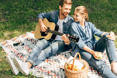 高角度的英俊的年轻人玩吉他为他的微笑的女朋友, 而放松在公园