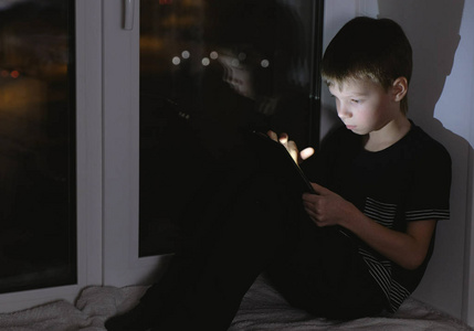 7year 老男孩在平板电脑上看东西。晚上在黑暗中坐在窗台上