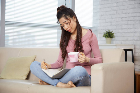 沉思的年轻妇女喝茶和阅读笔记在她的课本