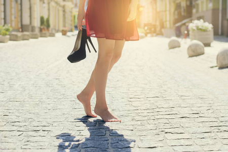 穿着红色高雅连衣裙的妇女, 手捧高跟鞋, 赤脚走在城里.关闭裁剪照片.
