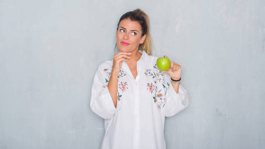 灰色垃圾墙上的年轻成年妇女吃新鲜的绿色苹果严肃的面孔思考问题, 非常迷惑想法