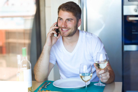 一张英俊的年轻人在家里的厨房里喝白酒时说的智能手机