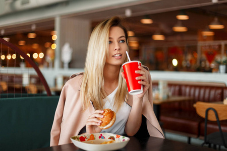 美丽的年轻模特女士穿着粉红色的夹克, 在咖啡馆里吃快餐, 喝一杯可乐。