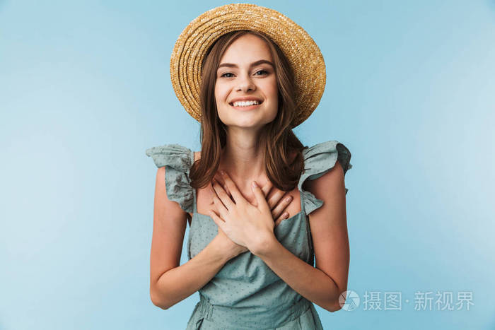 一个开朗的年轻女子在礼服和夏天的帽子摆出蓝色背景孤立的肖像