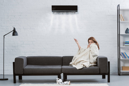 裹着毯子的年轻妇女在沙发下挂在墙上的空调和吹冷的空气