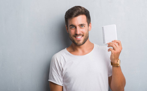 英俊的年轻人在灰色垃圾墙持有空白笔记本与一个快乐的脸站在微笑着自信的微笑显示牙齿