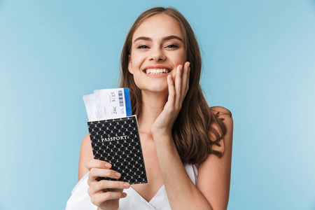 一个年轻的女孩在夏天的衣服戴着太阳镜的肖像持有护照与飞行门票隔离在蓝色背景