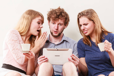 浏览朋友在平板电脑上冲浪互联网。年轻人，男人和女人在家里放松一下。女孩的家伙的耳朵低语