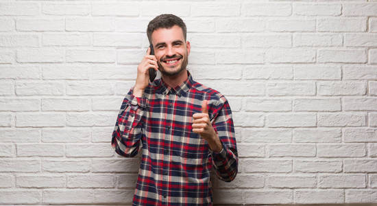 年轻人讲电话站在白色砖墙愉快与大微笑做 ok 标志, 拇指用手指, 优秀标志