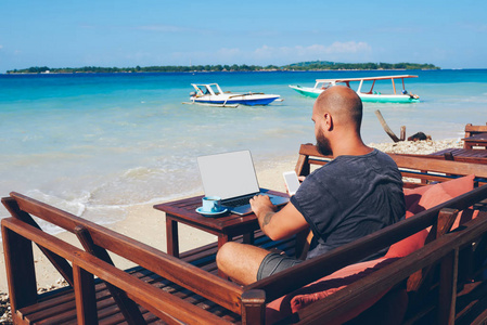 休闲服装的商人在岛上度假时使用上网本和无线互联网, 男性千禧年持有手机, 同时查看在海滩上的应用程序预订预订