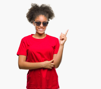 年轻的美国黑人妇女戴着太阳镜在孤立的背景下, 脸上带着微笑, 用手和手指指着侧面看着相机。