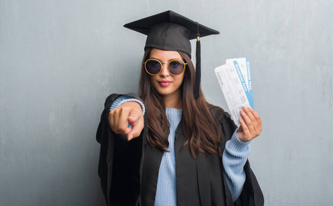 穿灰色墙壁的年轻黑发妇女穿着毕业生制服持有登机牌指向相机和你, 手势, 正面和自信的手势从前面