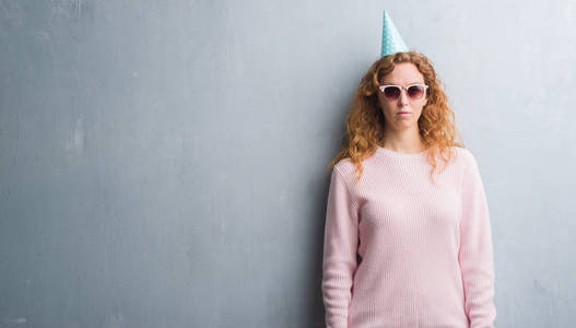灰色垃圾墙上的年轻红头发的女人戴着生日帽带着自信的表情对聪明的脸认真思考