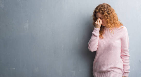 穿灰色垃圾墙的年轻红头发的妇女穿着粉红色毛衣疲倦的揉鼻子和眼睛感到疲倦和头疼。压力和挫折概念