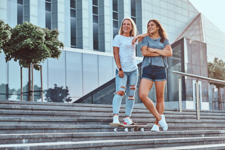 两个美丽的时髦女孩站在台阶上, 在摩天大楼的背景上的滑板
