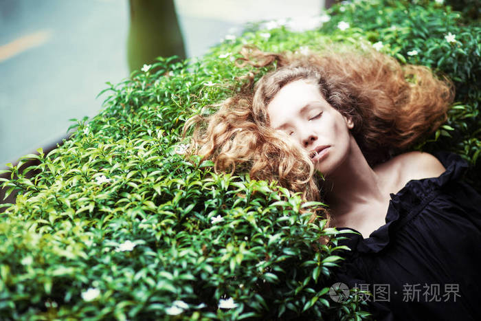 美丽的年轻红卷发妇女放松在灌木丛中的公园