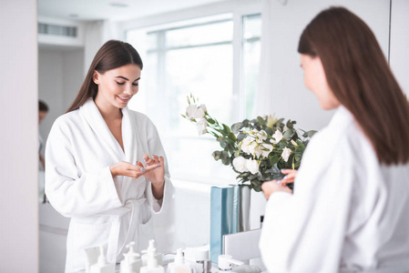 在白色明亮的公寓里, 在鲜花旁的浴袍上站在镜子旁边的快乐女人的肖像, 保持着护理皮肤的特殊液体
