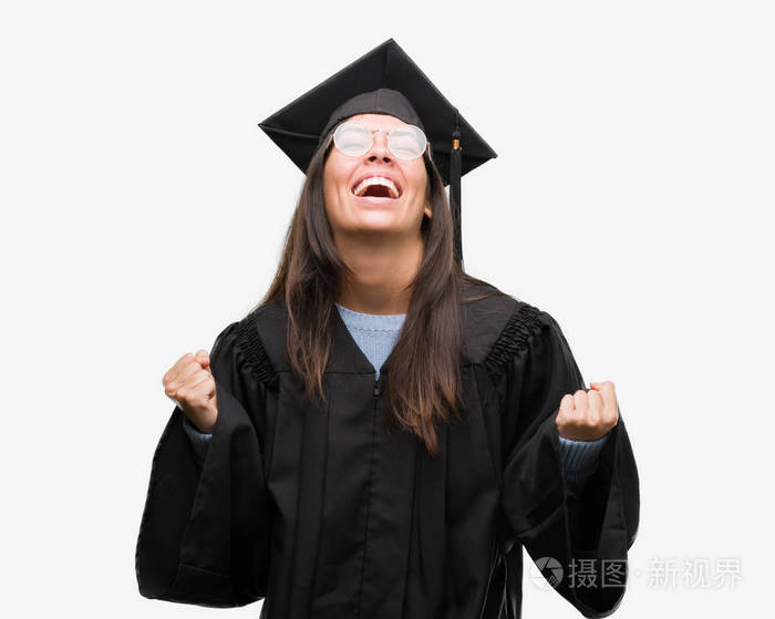 年轻的西班牙裔妇女穿着毕业帽和制服非常高兴和兴奋做赢家手势举起双臂, 微笑着, 尖叫着要成功。庆祝概念