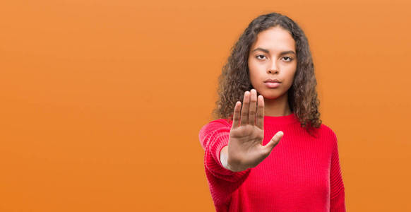 年轻的西班牙裔妇女穿着红色毛衣停止唱歌与手掌的手。脸上带有否定和严肃手势的警告表达式
