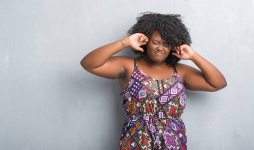 年轻的非洲裔美国妇女在灰色的垃圾墙上穿着五颜六色的礼服覆盖耳朵用手指与愤怒的表达大声音乐的噪音。聋的概念