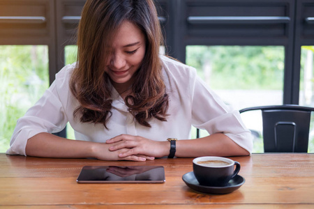 一个美丽的亚洲女人, 脸上带着笑脸看着绿色自然背景的现代咖啡馆木桌上的平板电脑
