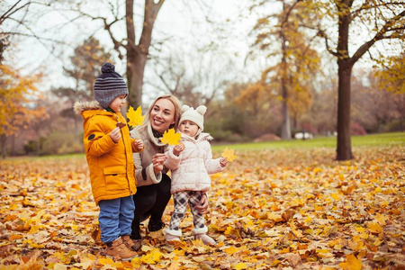一个年轻漂亮的母亲和两个小孩在秋季公园散步。妈妈和两个小孩玩耍。温暖的冬天。明亮的秋天