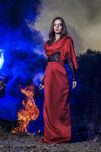美丽的时装模特女孩穿着红色的长裙和一条红色的披肩站在大火的背景下, 蓝色烟雾在日落。概念, 时尚, 现代和广告设计
