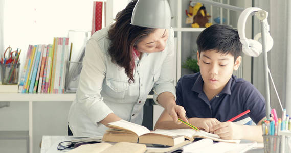 可爱的亚洲母亲帮助您的儿子在家做家庭作业与微笑面孔一起
