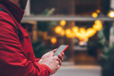 侧面裁剪视图的男性人在红色夹克触摸与现代智能手机设备的手指屏幕在夜间街道户外。手持手机的男子手拿着散景灯在复制空间附近