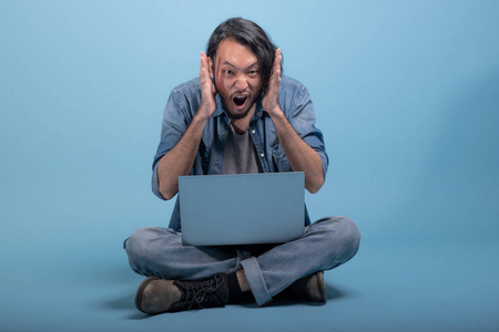 年轻的大胡子男子坐在地板上使用计算机在蓝色背景。疯狂的亚洲年轻的时髦使用膝上型电脑, 全身射击。年轻一代时髦的工作理念
