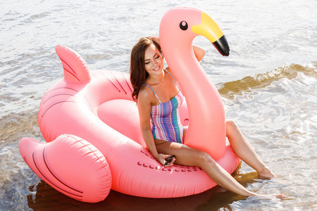 户外肖像的美丽的黑发模特在丰富多彩的夏季泳装。年轻美丽的女人坐在粉红色充气床垫形式的火烈鸟在白天, 波浪状的水背景