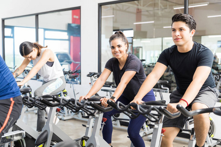 自信的男性客户微笑, 而在自行车上与朋友在健身房运动