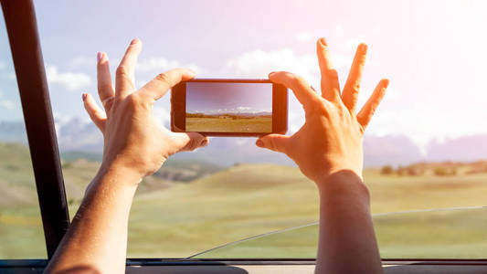 在汽车上, 一个年轻的游客特写照片, 在一个蓝色的天空背景下覆盖着积雪的山上的电话上拍照。旅游生活方式与生存